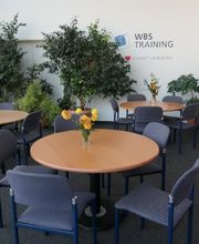 Gemütlicher Aufenthaltsraum mit runden Tischen, auf denen Blumen stehen, grünen Zimmerpflanzen, Bücherregal und rotem Sofa. Auf der Wand steht WBS TRAINING – unser Herz schlägt für Bildung geschrieben.