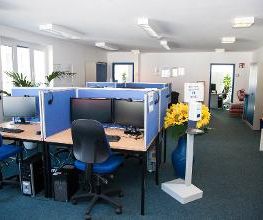 Dieses Bild zeigt einen Schulungsraum. Zu sehen sind Schreibtische, welche jeweils mit zwei Bildschirmen, einer Tastatur und Maus sowie einem Headset ausgestattet sind. In dem Raum befindet sich eine große Vase mit Sonnenblumen. 