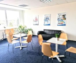 Aufenthaltsbereich kleine weiße Tische mit braunen Stühlen, Teppichboden und schwarzer Couch im Hintergrund