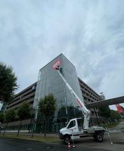 mediaService Bremen | Full-Stack Werbeagentur - Ein weißer Lkw mit einer ausgefahrenen Mateco-Arbeitsbühne montiert ein großes rotes Werbelogo an der Glasfassade eines modernen mehrstöckigen Gebäudes.