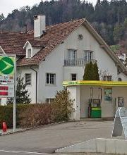 AGROLA Tankstelle in Zuckenriet (Niederhelfenschwil). Vor Wohnhaus Tankstelle mit kleiner Überdachung. Eine Tanksäule. Im Vordergrund Auffahrtstrasse mit Schild.