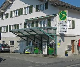 AGROLA Tankstelle in Einsiedeln. Mit kleiner Glasüberdachung. Zwei Tanksäulen und ein Zahlterminal. An Wohnhaus gelegen. Totem an Wohnhaus mit Preisanzeige und AGROLA Logo.
