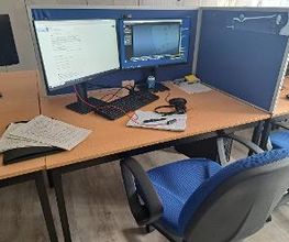 Arbeitsplatz mit 2 Bildschirmen, einer Maus, einer Tastatur und einem Headset. Auf dem Tisch liegen mehrere Dokumente.