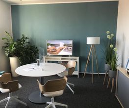 Aufenthaltsraum mit blauer Wand. Davor ein Regal mit TV. Daneben eine Stehlampe und mehrere Pflanzen. Im Raum ein Tisch mit 3 Stühlen