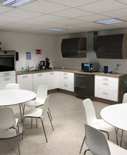 Küche mit 2 Tischen und jeweils 6 Stühlen. Küchenzeile mit Mikrowelle, Wasserkocher, Backofen, Dunstabzugshaube und Kaffeemaschine. Pflanzen stehen im Raum verteilt.