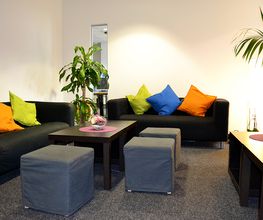 Gemütlicher Pausenraum mit 2 Sofa auf denen bunte Kissen liegen, 2 Tischen und drei Höckern. Außerdem befinden sich mehrere grüne Zimmerpflanzen im Raum.