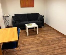 Eine schwarze Couch in einem Pausenraum. Am Bildrand ist ein Holztisch mit einem weiteren Stuhl zu sehen.