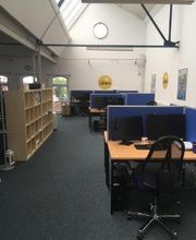 Schulungsraum mit mehreren Arbeitsplätzen mit jeweils 2 Bildschirmen, einem Bürostuhl, Tastatur, Maus und Headset. Links steht ein Holzregal.