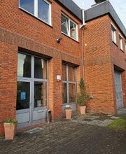 Ein Backsteingebäude mit großen Fenstern und einer Glastür. Vor dem Eingang stehen Blumenkübel mit Pflanzen. Rechts ist ein großes Garagentor zu sehen. Der Boden vor dem Gebäude besteht aus Pflastersteinen und einem kleinen Bereich mit Gras.