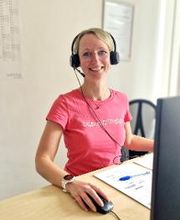 Eine Mitarbeiterin sitzt an ihrem Schreibtisch und arbeitet an ihrem Computer. Sie trägt ein Headset und lächelt freundlich in die Kamera.
