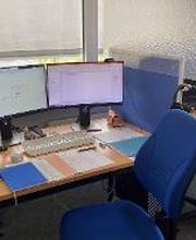 Arbeitsplatz mit 2 Bildschirmen, einem Bürostuhl, Tastatur, Maus und Headset. Ein Block sowie 2 Trinkflaschen stehen auf dem Tisch.