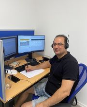 Ein Teilnehmer sitzt an seinem Arbeitsplatz vor 2 PC-Bildschirm und trägt sein Headset. Er macht sich Notizen und schaut gut gelaunt in die Kamera.