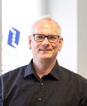 Auf dem Bild ist Ulrich Jung, Regionalleiter der WBS TRAINING, zu sehen. Er trägt ein schwarzes Hemd und lächelt freundlich in die Kamera. Im Hintergrund ist ein Fenster zu erkennen, das einen Teil des Firmenlogos der WBS TRAINING zeigt