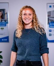 Bild einer Mitarbeiterin. Blonde, gelockte Haare, blauer Pullover, freundliches Lächeln.