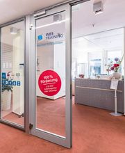 Eingangstür aus Glas mit Blick auf den Empfangstresen. Auf der Tür klebt ein roter Aufkleber auf dem steht 100 % Förderung sowie ein blauer Schriftzug von WBS TRAINING plus Logo. 