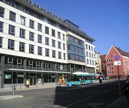 Ein vierstöckiges, weißes Gebäude an einer Straße. Vor dem Gebäude hält gerade ein Linienbus.