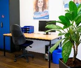Arbeitsplatz mit Fachablage und Bildschirm sowie Bürostuhl. darüber Plakate. Grüne Zimmerpflanze und blauer Schrank.