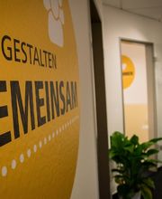 Eingangsbereich. Glastüre mit WBS TRAINING Aufschrift und Logo. Zimmerpflanze. Gelbes Wandtattoo mit der Aufschrift „Wir gestalten gemeinsam“.