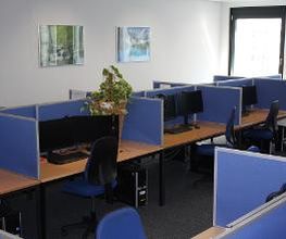 Ein Schulungsraum mit mehreren Lernplätzen, die durch blaue Trennwände voneinander getrennt sind. Jeder Arbeitsplatz besteht aus 2 PC-Bildschirmen, Tastatur und Maus sowie Headset.