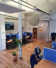 Schulungsraum mit mehreren Arbeitsplätzen jeweils mit 2 Bildschirmen, einem Bürostuhl, Tastatur und Maus. Im Hintergrund ein Regal neben der Tür. Mittig im Raum eine Säule und eine Topfpflanze.