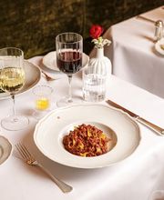 Refined restaurant table setting with tagliatelle in a rich meat sauce, crusty bread on side plates, red and white wine glasses, candlelight, and crisp white linens against a plush banquette backdrop.