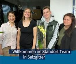 Team aus 4 Personen die freundlich in die Kamera schauen. Ein Kollege hält eine gelbe Orchidee, am unteren Bildrand der Text "Willkommen im Standort Team in Salzgitter"
