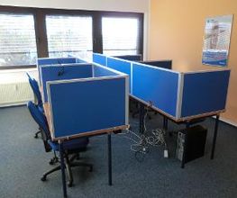 Schulungsraum mit 6 Arbeitsplätzen mit blauen Trennwänden. An der orangenen Wand hängt ein WBS TRAINING Plakat.