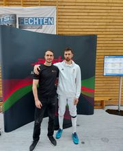 Zwei Personen stehen nebeneinander vor einer Veranstaltungswand im sportlichen Umfeld. Die Pose vermittelt Zusammenhalt und einen offiziellen Anlass.