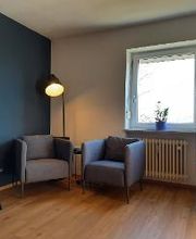 Aufenthaltsraum mit 2 Sesseln vor einer Stehlampe, daneben einem Regal mit Deko und einem weiteren Tisch mit Stuhl