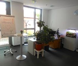 Ein Arbeitsplatz vor einer Fensterfront. Neben dem Tisch steht ein Flipchart mit der Aufschrift "Herzlich Willkommen!", auf der anderen Seite des Tisches steht ein beleuchtetes Aquarium.