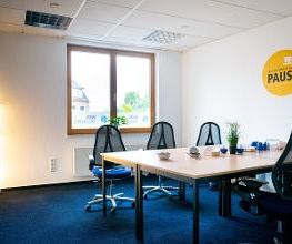 Pausenraum mit großem Tisch und mehreren Arbeitsplätzen und Bürostühlen. In der Ecke eine Stehlampe, an der Wand ein Sticker mit Aufschrift: Mach einfach mal Pause.