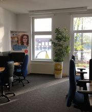 Schulungsraum mit mehreren Arbeitsbereichen, die durch blaue Trennwände voneinander abgegrenzt sind und mit 2 PC-Bildschirmen, Tastatur, Maus und Headset ausgestattet sind. Zugang zum Balkon.