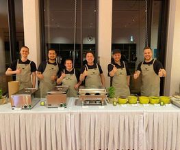 dean&david Catering | Stuttgart - Freundliches Catering Team hinter einer Buffetstation mit warmen Speisen, Saucen und frischen Kräutern für eine professionelle Eventbewirtung.