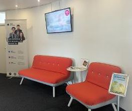 Wartebereich im Flur mit 2 modernen, roten Sesseln, einem dazugehörigen Beistelltisch, einem Fernseher an der Wand und einem Werbeaufsteller im Hintergrund.