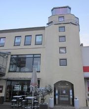 Außenansicht des Gebäudes, im Vordergrund Tische und Stühle, im Hintergrund blauer Himmel. 