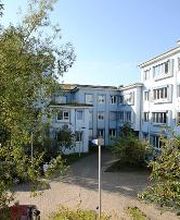 Ein großes Gebäude mit weißer Fassade und hellblauen Fensterrahmen. Es ist ein sonniger Tag, der Himmel ist blau und es sind grüne Baumkronen im Bild.