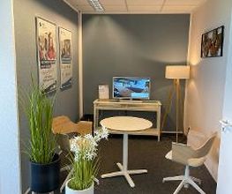 Beratungsbereich mit Tisch und 2 Stühlen. Im Hintergrund ein Regal mit TV, links stehen 2 Pflanzen, an der Wand hängen 2 Plakate.
