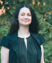 Frau mit schwarzem Haar steht vor grünem Baum und lächelt.