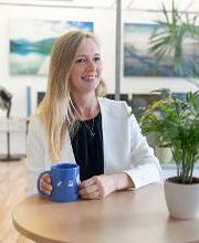 Frau mit blondem Haar steht am Tisch und hält eine blaue Tasse. Auf dem Tisch steht eine Topfpflanze. Im Hintergrund sieht man 2 Landschaftsbilder an der Wand und eine Tür auf der linken Seite.