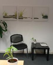 Stuhl mit Beistelltisch neben einer Couch und Topfpflanze. Im Hintergrund hängen drei Leinwandbilder von grauen Steinen und Gras an der Wand.