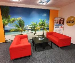 Pausenbereich bestehend aus 2 roten Sofas mit einem kleinen schwarzen Tisch. Großes Wandbild von einem Strand mit Palmen.