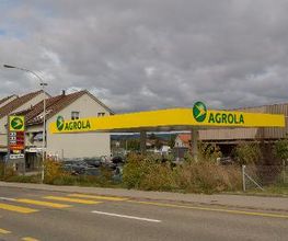 AGROLA Tankstelle in Rickenbach Sulz; diesige Aufnahme; vorne Strasse mit Fussgängerstreifen; links Totem mit AGROLA Logo und digitaler Preisanzeige; dahinter Wohnhäuser; rechts Tankstellendach