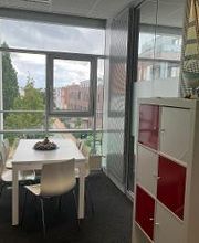 Ein heller Pausenraum mit einem weißen Tisch und vier weißen Stühlen. Rechts steht ein Regal mit roten und weißen Fächern, darauf ein Modell eines Segelboots. Große Fenster bieten einen Blick auf Bäume und umliegende Gebäude.