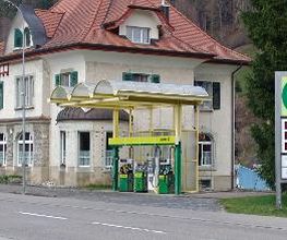 AGROLA Tankstelle in Brunnadern an Dorfstrasse gelegen. Rechts Totem mit AGROLA Logo und darunter digitaler Preisanzeige. Geschwungene Glasüberdachung der Tankstelle mit drei Tanksäulen