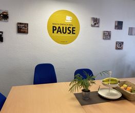 Ein Pausenbereich mit Tisch und Stühlen. Auf dem Tisch ein Obstkorb und eine Topfpflanze. An der Wand mehrere Kaffeebilder.