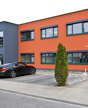 Außenansicht eines orange-grauen Gebäudes, im Vordergrund Parkplatz mit einem schwarzen PKW und ein Baum.