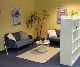 Pausenraum mit 2 Sofas, einem Beistelltisch mit Blumen und einem Regal als Raumtrenner mit Pflanzen. An der Wand 2 Plakate und ein Teppich auf dem Boden.