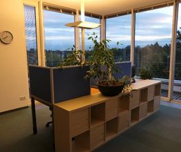 2 Arbeitsplätze bestehend aus 2 PC-Bildschirmen, Tastatur, Maus und Headset sowie blauen Trennwänden. Regal mit Zimmerpflanze. Im Hintergrund große, bodentiefe Fenster mit Blick auf den Balkon oder Dachterrasse.