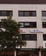Ein mehrstöckiges weißes Gebäude mit dunklen Fenstern. An der Fassade sind Schilder von „ver.di“ und „WBS Training“ angebracht. Im Erdgeschoss befindet sich eine Bäckerei mit dem Schild „Frische Backwaren“. Vor dem Gebäude steht ein großer Baum.