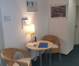 Eine kleine Sitzecke bestehend aus 2 gemütlichen Stühlen, einem Tisch und einer kleinen Lampe. Auf dem Tisch liegt Informationsmaterial und an der Wand hängen einige Bilder.